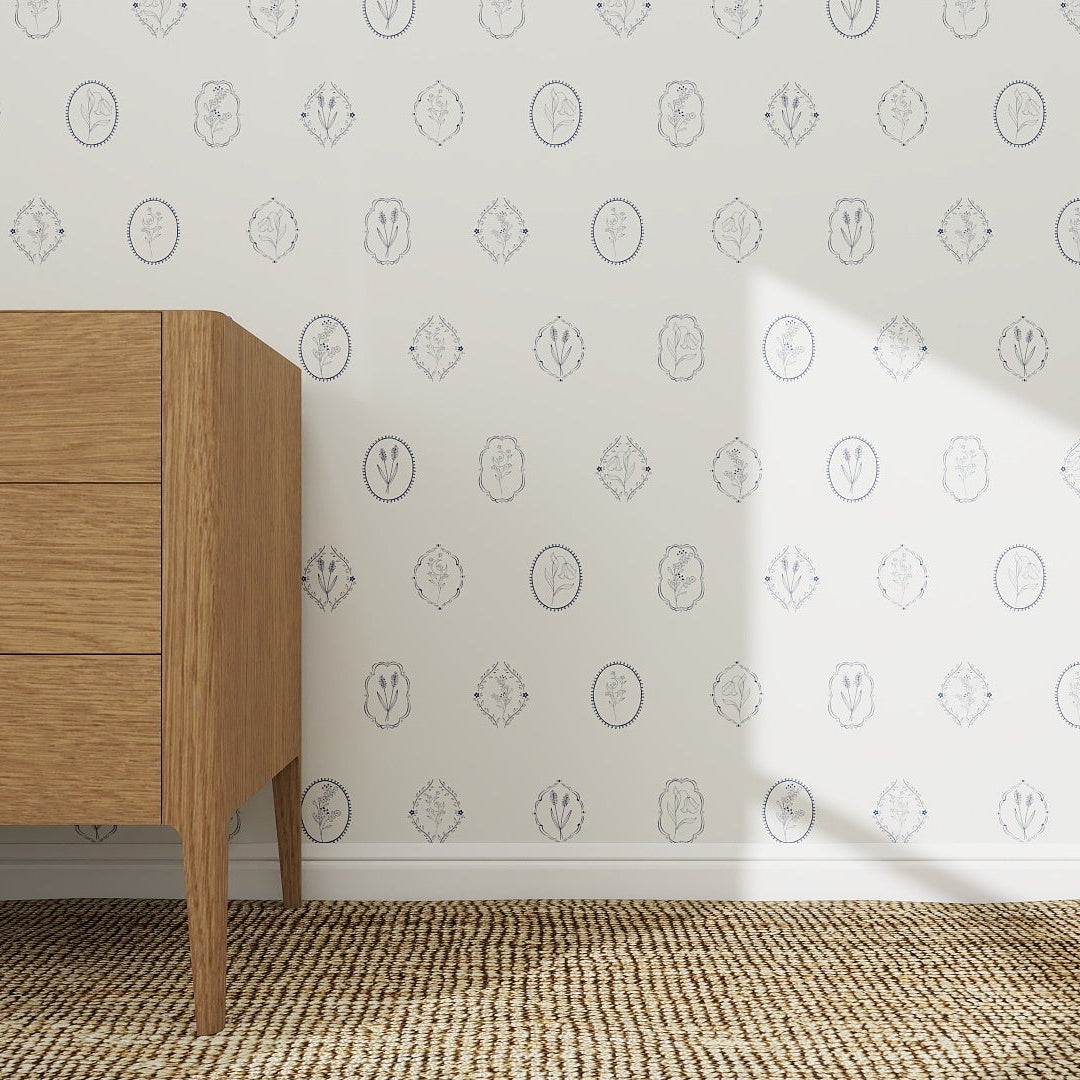 A wooden dresser sits on a woven rug beside a white wall adorned with Botanic Emblem Wallpaper | Sample, featuring blue outlined leaves and animal faces. Sunlight casts gentle shadows on the wall and floor.