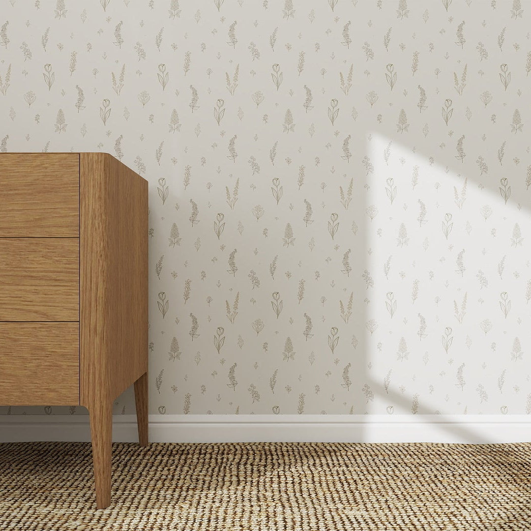 A wooden dresser with three drawers stands on a woven rug by a white wall decorated with Delicate Meadows Wallpaper | Sample, featuring a subtle beige floral pattern. Sunlight casts a diagonal shadow, enhancing the décor’s colour and quality.