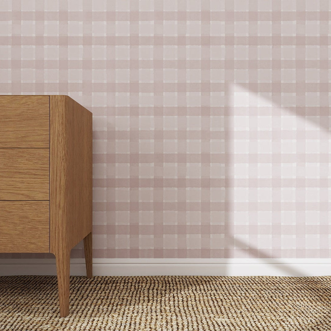 A wooden dresser with three drawers sits on a woven rug beside a wall covered in Checkered Wallpaper Repeat Pattern Pink | Sample. Sunlight casts a shadow, highlighting the quality of the wallpaper samples pink checks.