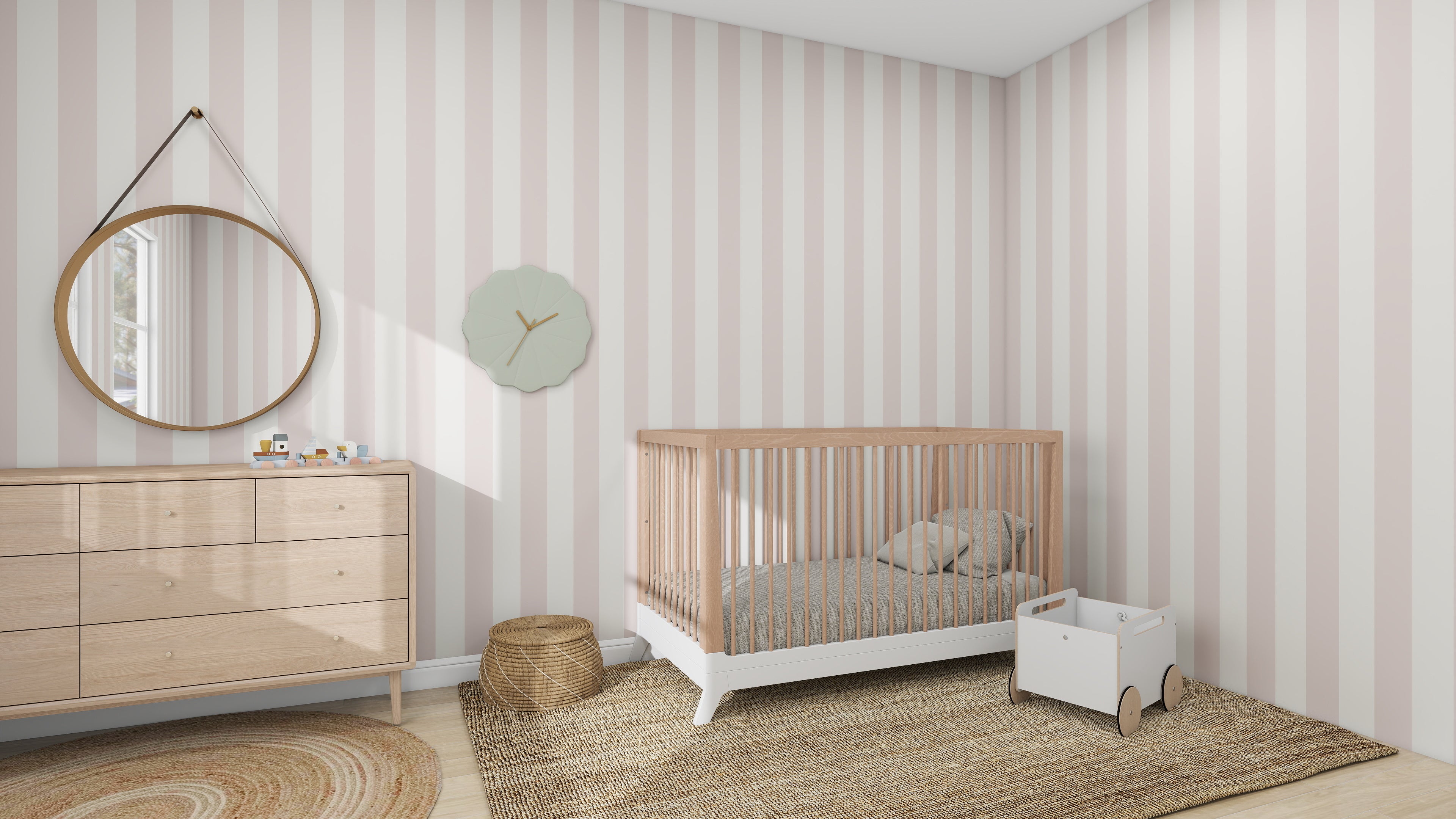 In this inviting nursery, the munks and me Circus Stripe Wallpaper in Blush Rose sets a playful tone. A wooden crib and light wood dresser with toys and mirror add charm. Nearby, a small round woven basket sits beside a toy cart, and a minimalist clock hangs above the crib.