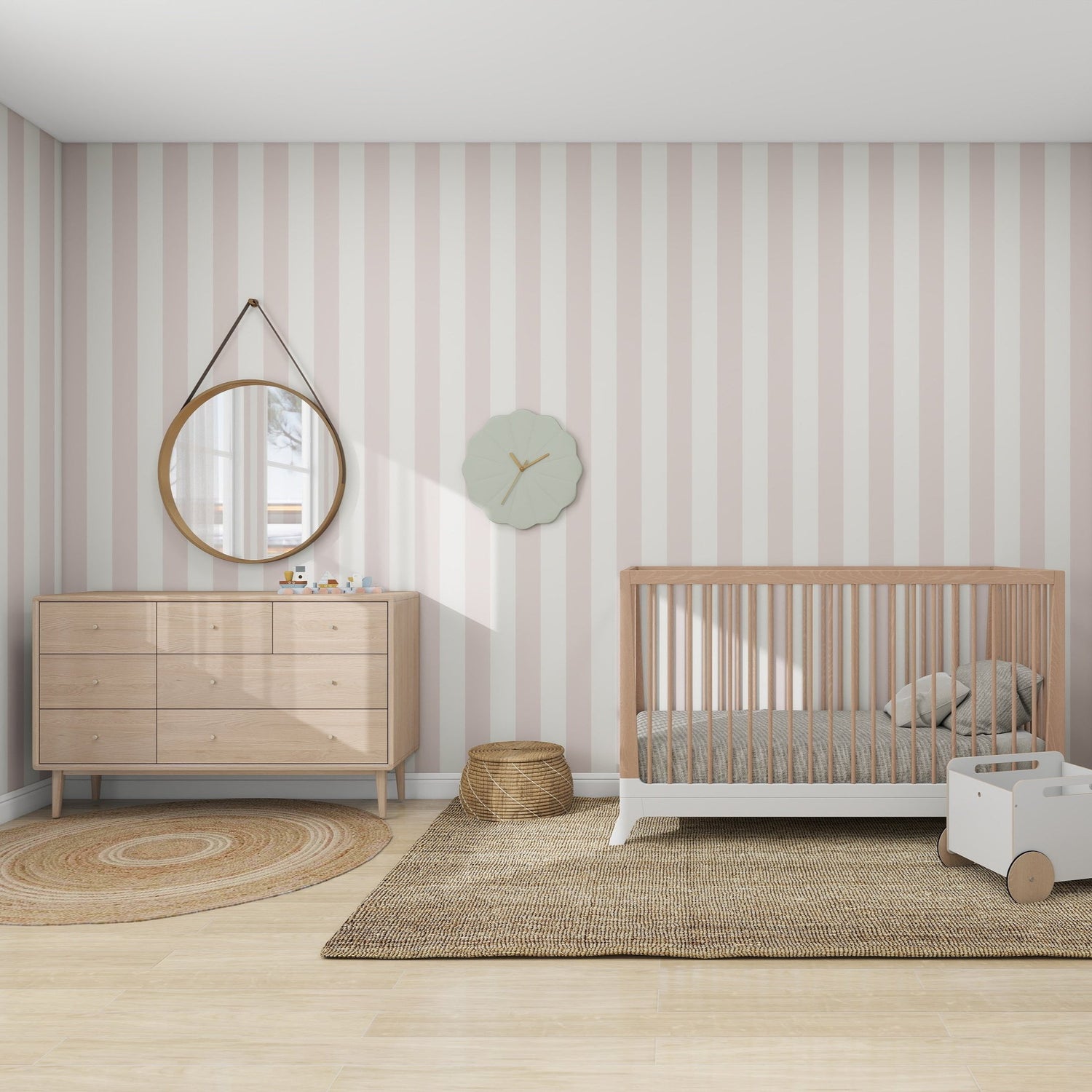 A cozy kids space with munks and mes Circus Stripe Wallpaper Blush Rose features a wooden crib, a dresser with a round mirror above, a round woven rug, small basket, decorative wall clock, white toy cart, all on an expansive beige rug.