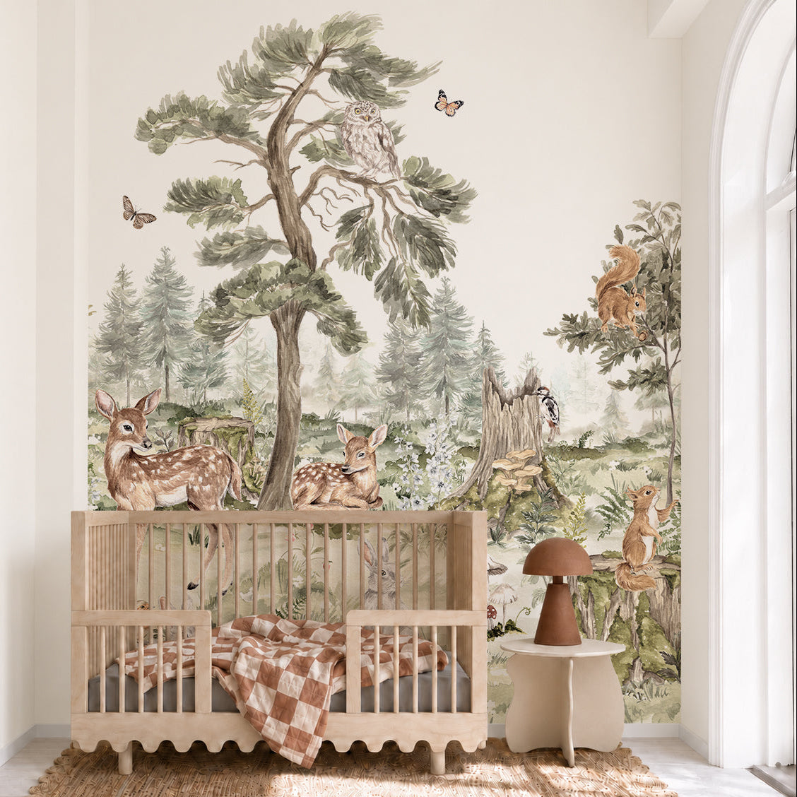 A cozy nursery features the “Into The Forest Mural,” a hand-painted woodland scene with deer, owls, squirrels, rabbits, butterflies, and trees behind a wooden crib. Natural light pours through a large arched window, illuminating the enchanting nature-inspired wall decor.