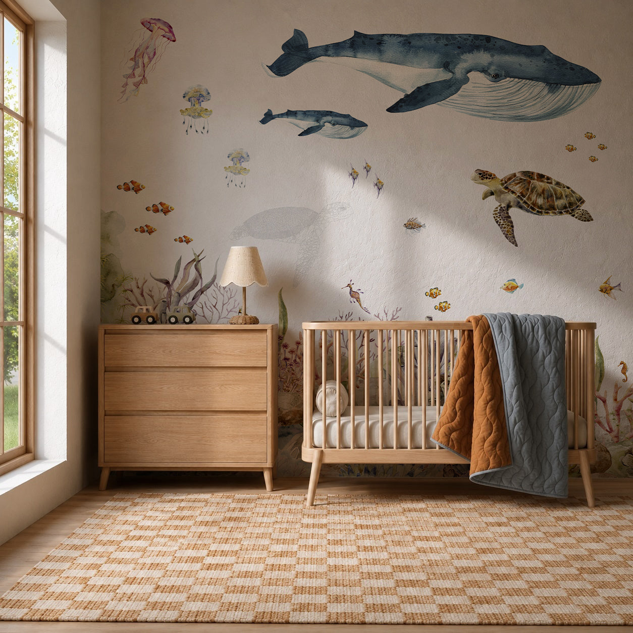 A cozy nursery with a wooden crib and dresser features the Under The Sea Mural, Watercolour, adorned with whales, fish, and a turtle. Ocean-themed wall art complements the playful sea creatures of the mural, while a lamp and large window let in natural light. A quilt is draped over the crib.