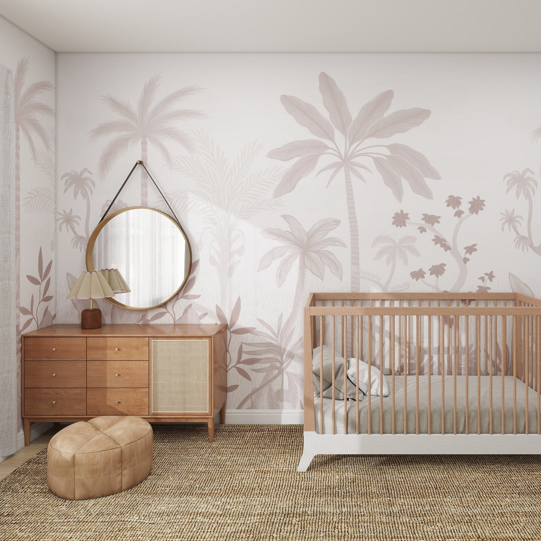 A cozy nursery includes a wooden crib, brown dresser with a round mirror, small lamp, and light tropical palm walls using NATURAL WONDER PALM WALLPAPER MURAL ROSE. A woven rug adorns the wooden floor. Nearby, a colorful pouf adds vibrancy by the open door.