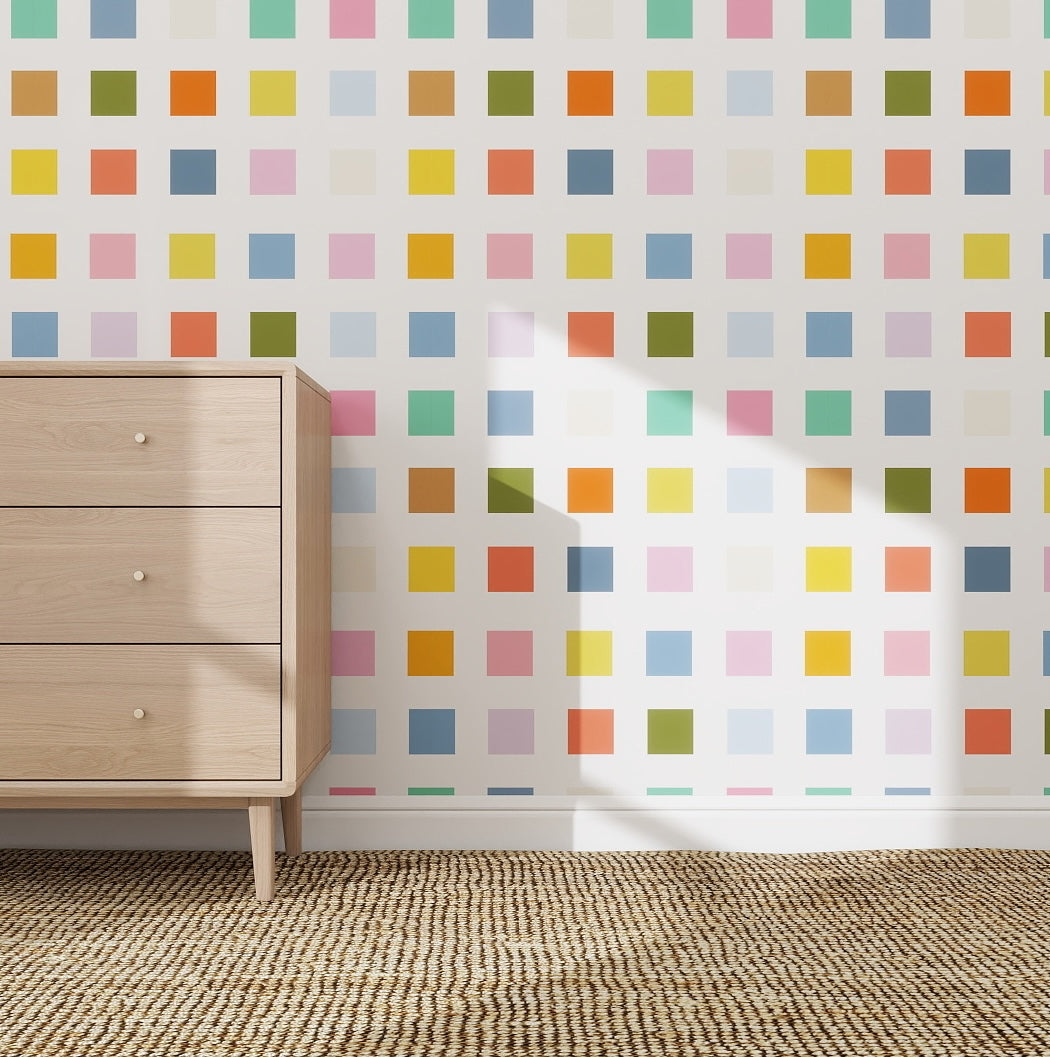 Rainbow Squares Wallpaper Repeat Pattern adds color and creativity to your space. Here, it decorates a wall beside a light wood dresser on a woven rug, while sunlight highlights the pastel squares’ playful design.