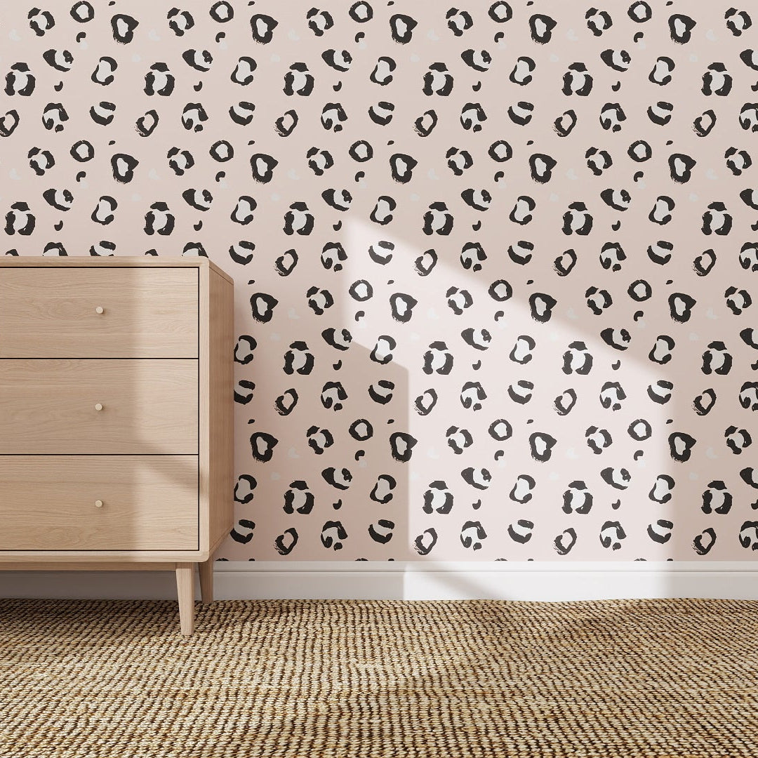 A light wooden dresser sits on a woven rug before a wall decorated with Leopard Print Wallpaper Repeat Pattern | Pink, as sunlight streams in and playful shadows highlight the rooms quirky style.