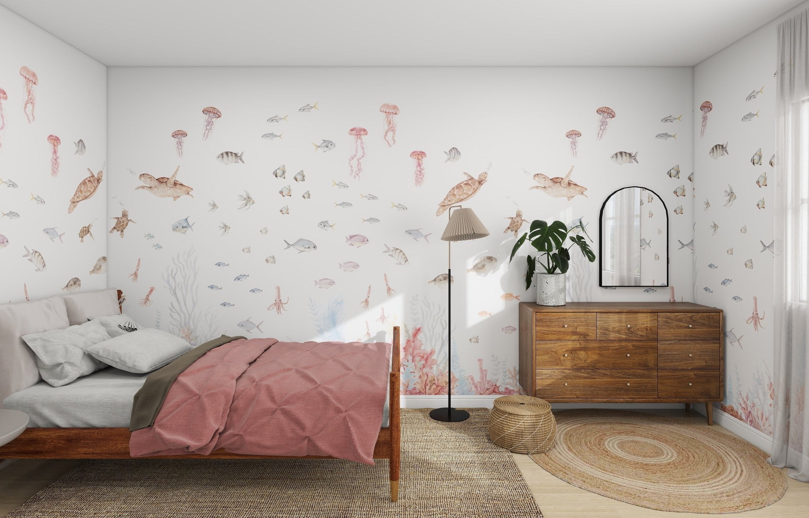 The bedroom showcases the Fish Tank Wallpaper Mural by munks and me, depicting sea life with fish, turtles, and jellyfish. On the left sits a pink-dressed wooden bed; on the right, a rustic dresser with a mirror and plant. A lamp stands nearby on a woven rug enhancing the rooms aquatic vibe.