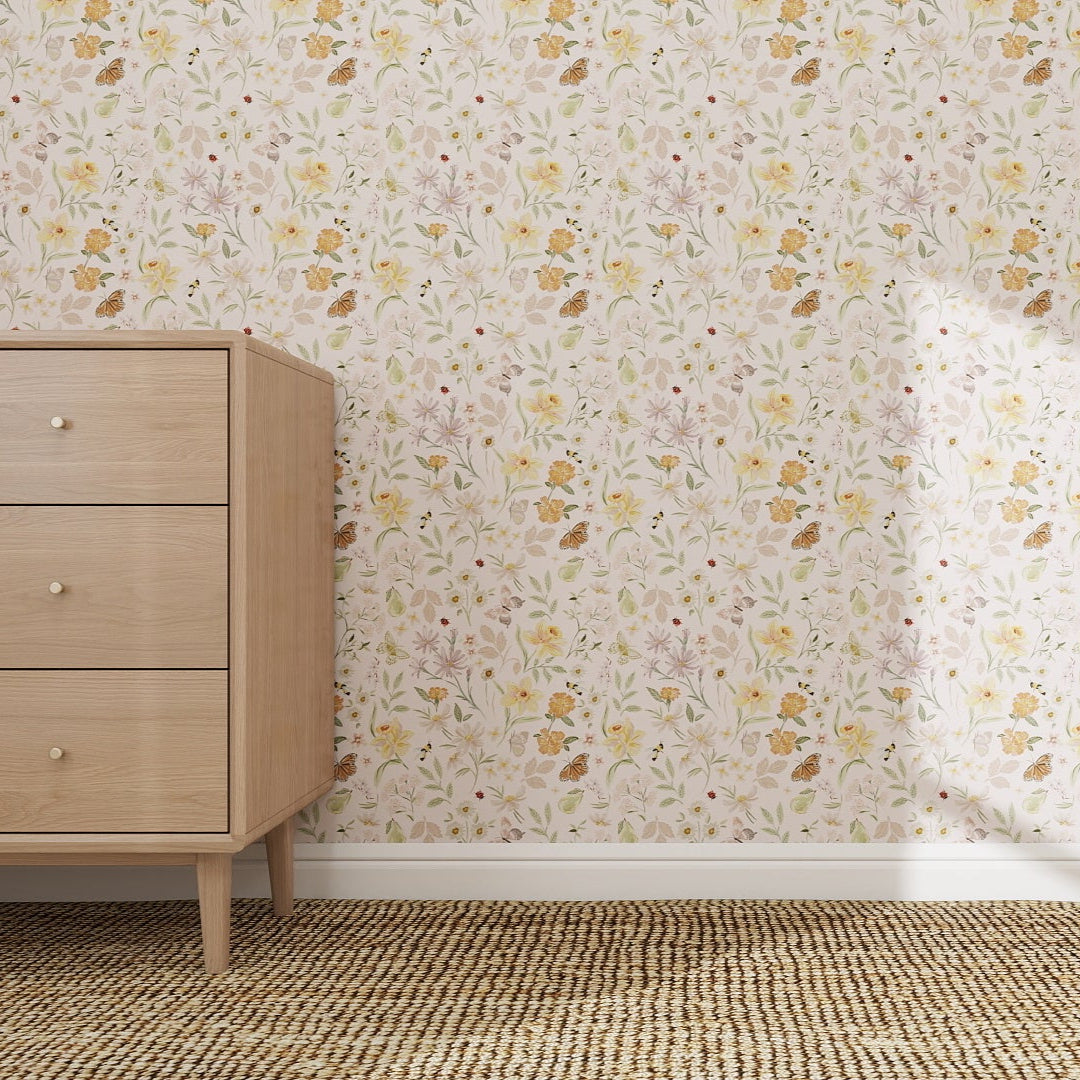 A wooden dresser with three drawers stands on a woven rug beside a wall featuring the Garden Symphony Wallpaper Repeat Pattern | Sample in soft pastel florals. The scene is brightly lit and minimalist.