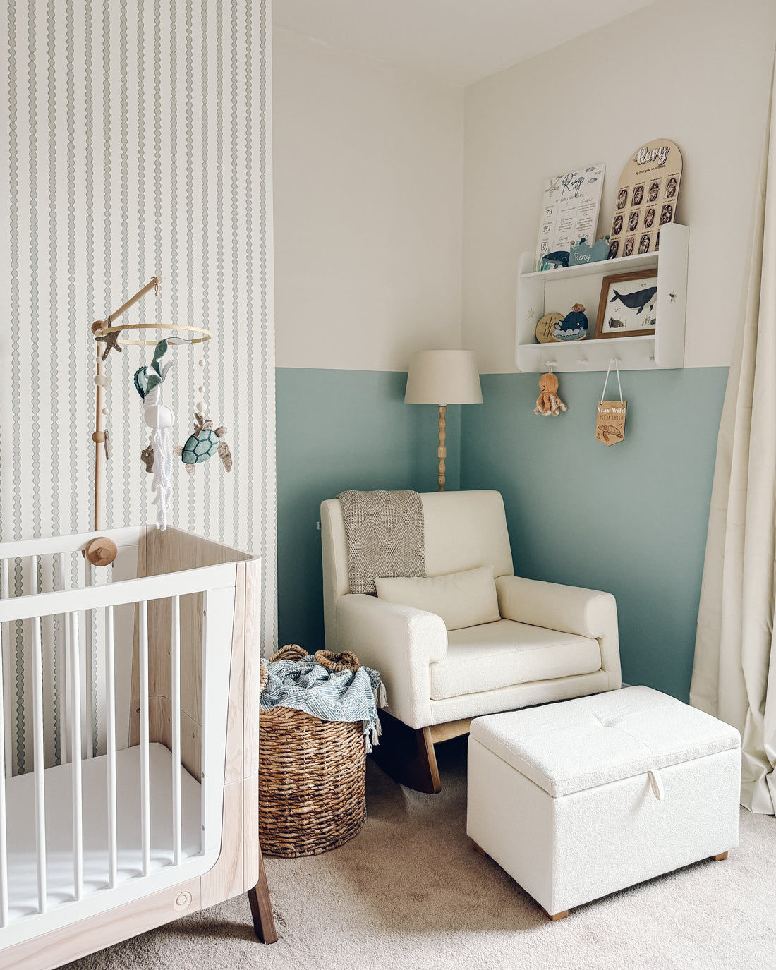 A cozy nursery features a white crib, a wicker basket, comfy armchair and ottoman, plus a floor lamp. Little Abacus Stripe Blue and Green wallpaper pairs perfectly with half-painted teal and white walls for added charm.