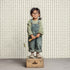 A young child in glasses, a green checked shirt, and denim overalls stands on a wooden box with a paint roller, set against vibrant Jolly Jester Wallpaper Repeat Sage Green.