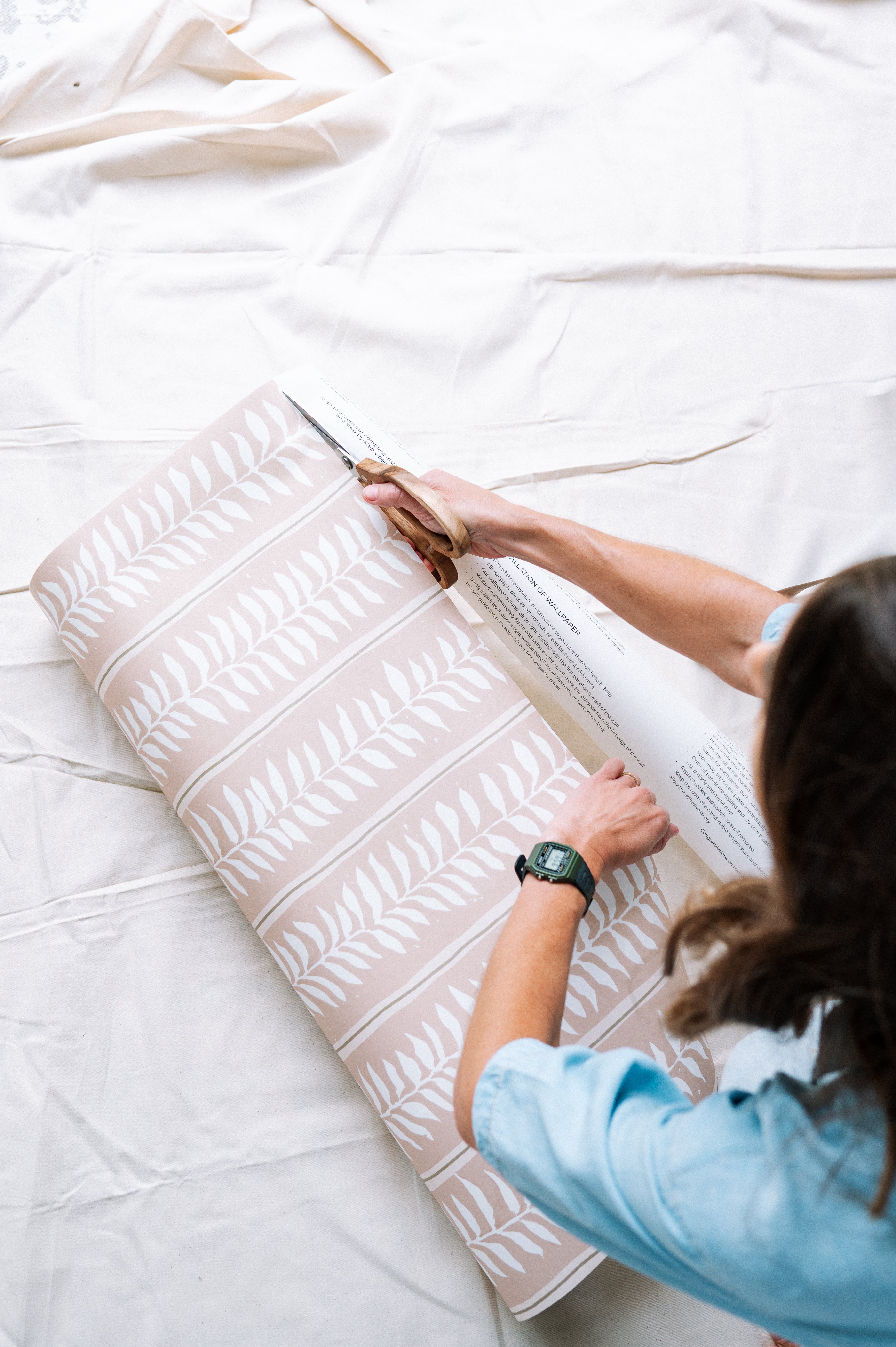 A person in a blue shirt and watch cuts a roll of Meadow Leaf Stripe Wallpaper, Clay, featuring pink botanical designs with white leaf patterns, on a white surface.