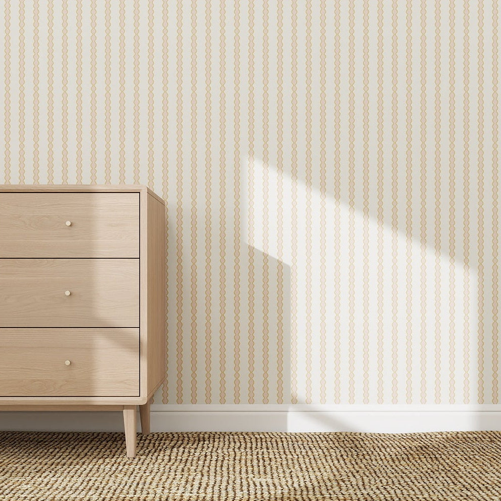 A light wood dresser with three drawers stands on a patterned beige carpet, against a wall decorated with Little Abacus Stripe Wallpaper Sample in pink and yellow. Sunlight casts a shadow across the wall and floor.