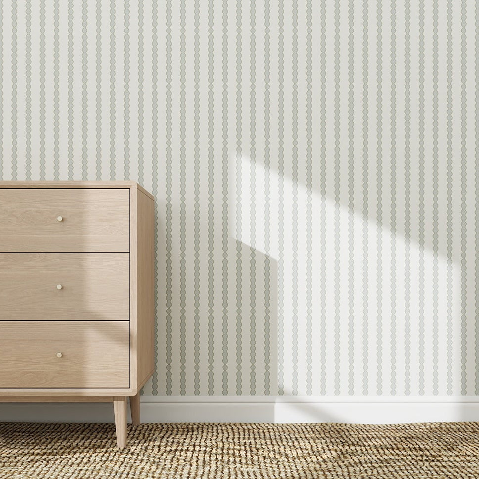 A light wood dresser with three drawers stands on a beige patterned rug before a wall covered with Little Abacus Stripe Wallpaper Sample in blue and green. Sunlight casts a bright diagonal shadow across the striped wall.