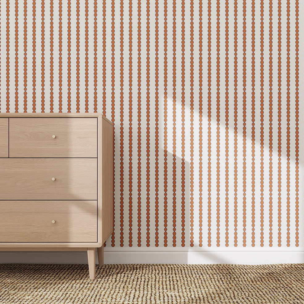 A light wood dresser with three drawers sits on a woven rug before a white wall featuring Little Abacus Stripe Wallpaper, Red and Blue. Sunlight casts soft shadows, highlighting this playful kids room wallpaper’s modern style.