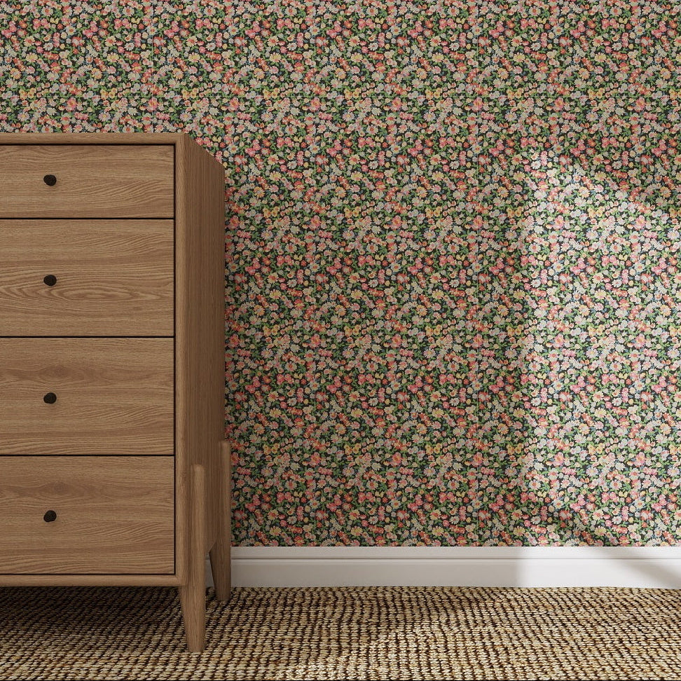 A wooden dresser stands on a woven rug beside a wall adorned with Midnight Meadow Florals Wallpaper Sample—sunlight highlights its delicate pink, green, and yellow pattern.