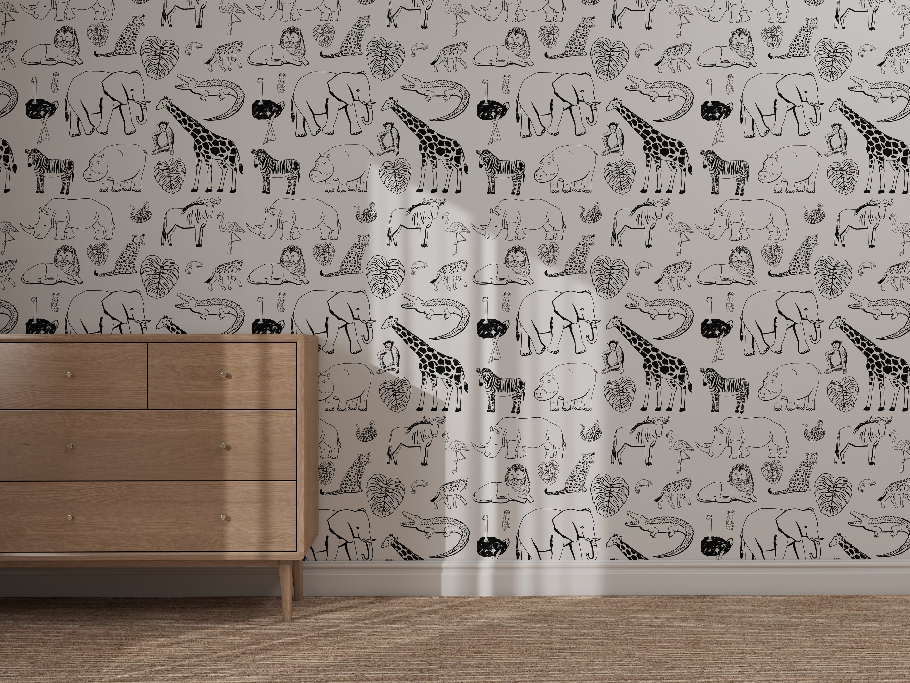 A light wooden dresser stands before the Going on Safari Wallpaper Repeat Pattern | Black and White, showcasing black-and-white giraffes, rhinos, elephants, leopards, and leaves as sunlight casts soft shadows on the illustrations.