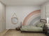 A cozy childrens space featuring a single bed with sage green bedding, a round mirror, woven rug, basket, small dark nightstand with lamp, and the Corner Rainbow Wallpaper Mural as a pastel feature wall.
