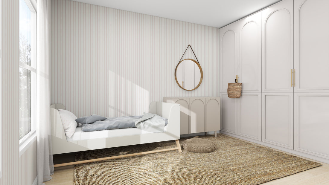 A minimalist bedroom features a small white bed with gray bedding and a round mirror above a cabinet. A woven basket adds texture, while the walls are adorned in Pinstripe Wallpaper Blush Rose by munks and me. Natural light floods through the large window, enhancing the neutral palette.