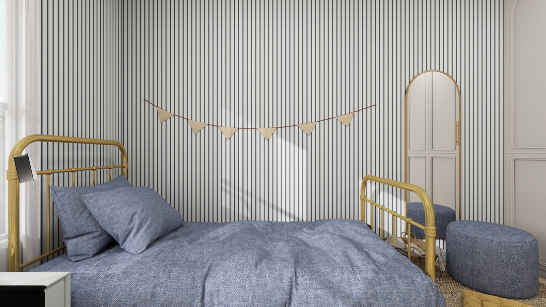 The room showcases the Pinstripe Wallpaper Denim sample by munks and me, paired with a cozy bed draped in a blue duvet. Gold-toned bed frame accents enhance the elegance. A tall mirror and round blue ottoman sit nearby under stringed pennant flags.