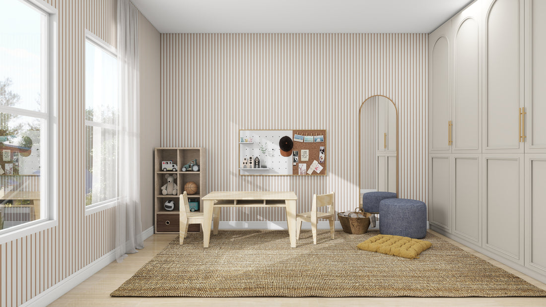 A cozy playroom with Pinstripe Wallpaper Latte from munks and me features a wooden table with two chairs on a woven rug. A bulletin board displays colorful art, while toy-filled shelves, two blue poufs, and a tall mirror add charm as light streams through large windows.