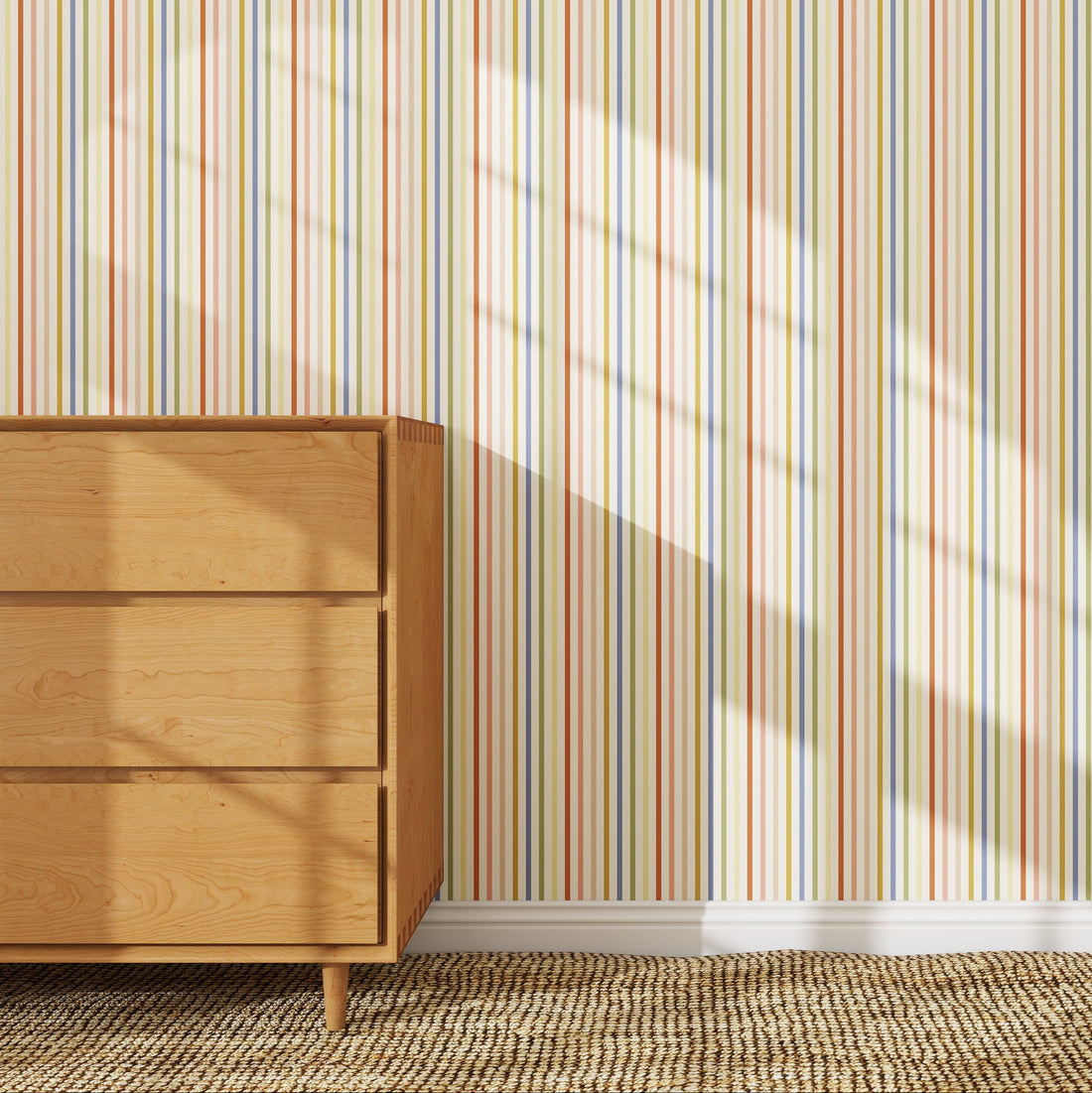A wooden dresser stands on a woven rug against a wall covered with Pico Stripe Wallpaper Sample, Multi. Sunlight streams in, casting patterned window shadows across the high-quality striped wallpaper and dresser.