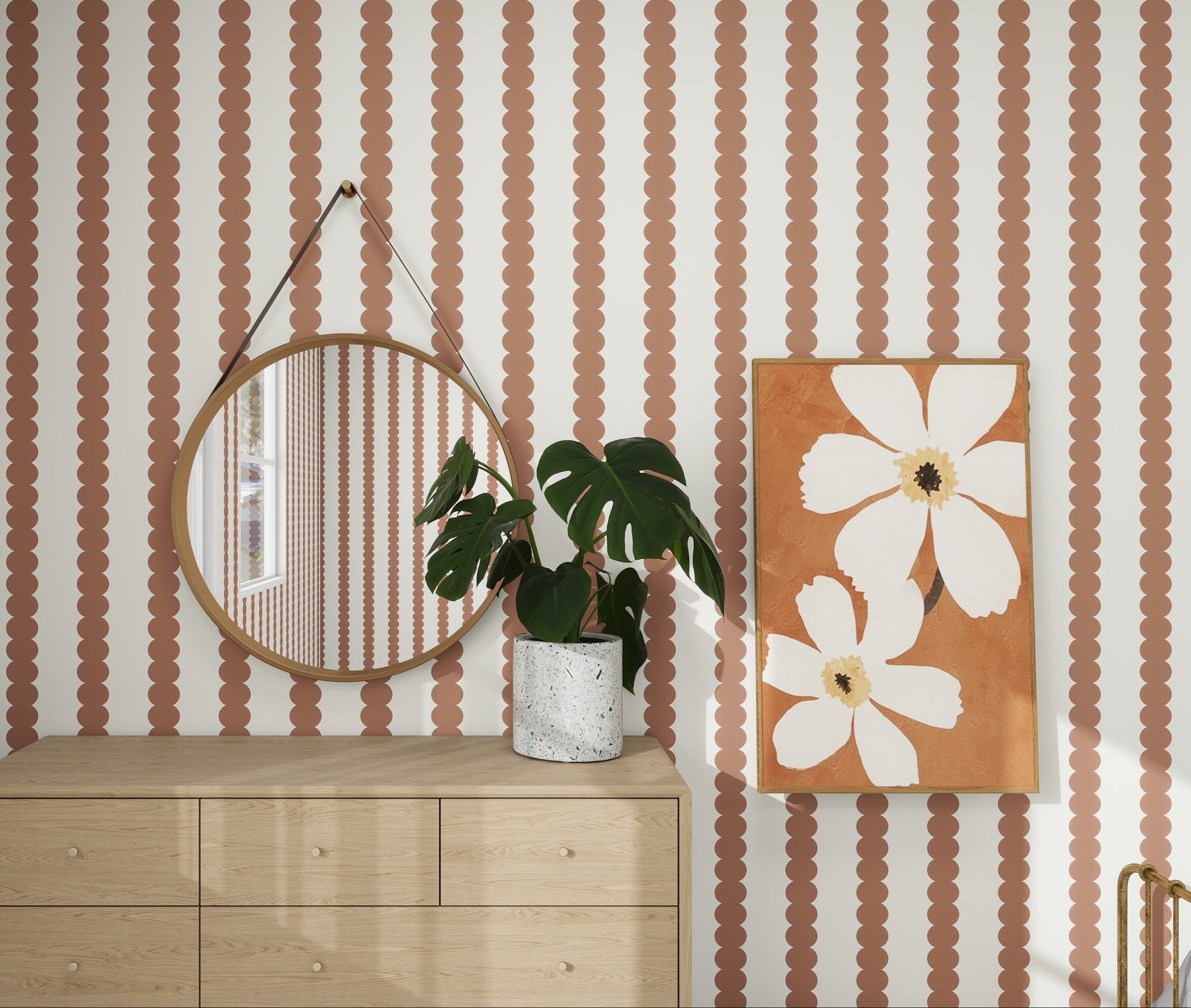 A nursery features the Mocha-colored dresser with a circular mirror reflecting munks and mes Scallop Stripe Wallpaper in Mocha. A potted plant sits on the dresser, and to the right, a framed artwork of white flowers on an orange background adds whimsy.