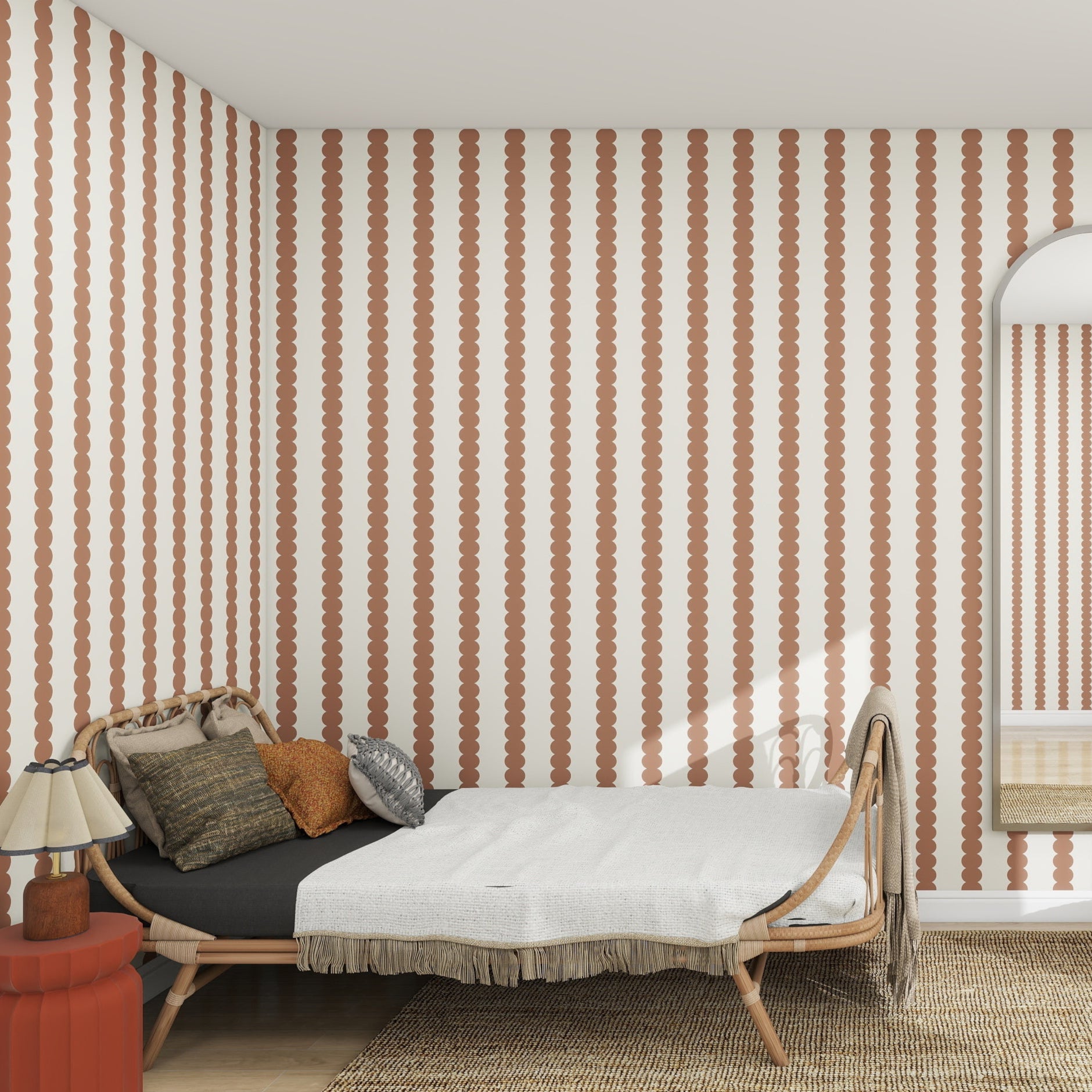 In a cozy bedroom, a bed with a white blanket and decorative pillows sits against a wall adorned with munks and mes Scallop Stripe Wallpaper in Mocha. A red side table with a fringed-lamp adds warmth, while a large mirror leans on the wall, enhancing the inviting ambiance.