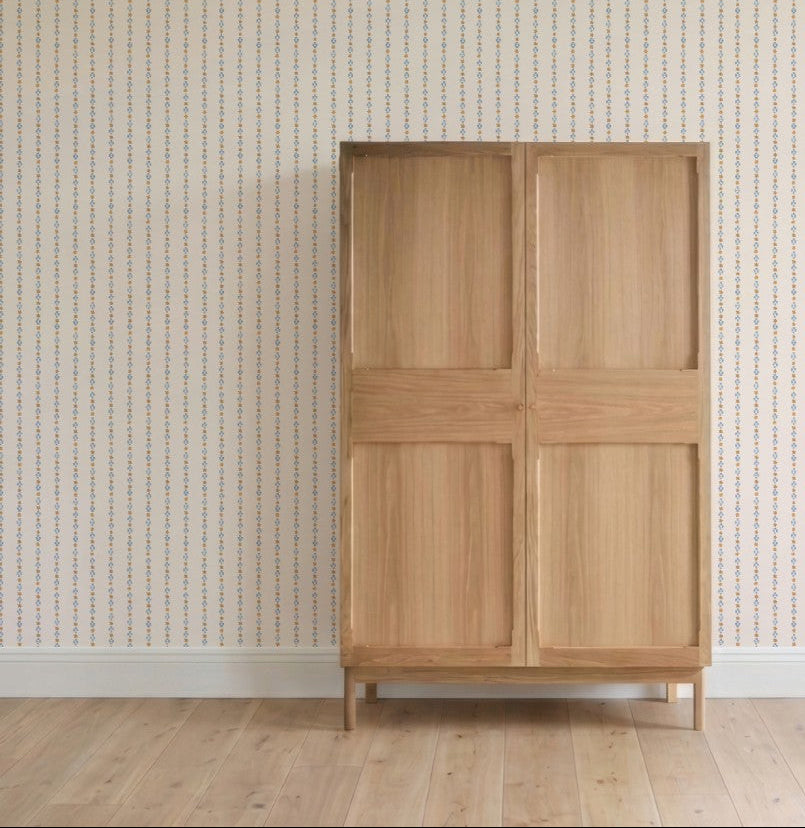 A simple wooden wardrobe with four panel doors stands against a wall adorned with Jacquard Stars Wallpaper in gold. Light wood flooring and white baseboards complete the room&
