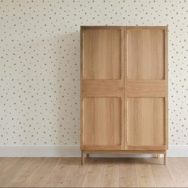A light wooden wardrobe with two doors stands on a light wood floor, backed by a wall featuring Soaring Hearts Wallpaper Sample—its colorful floral pattern highlights excellent quality in this minimalistic, tidy room.