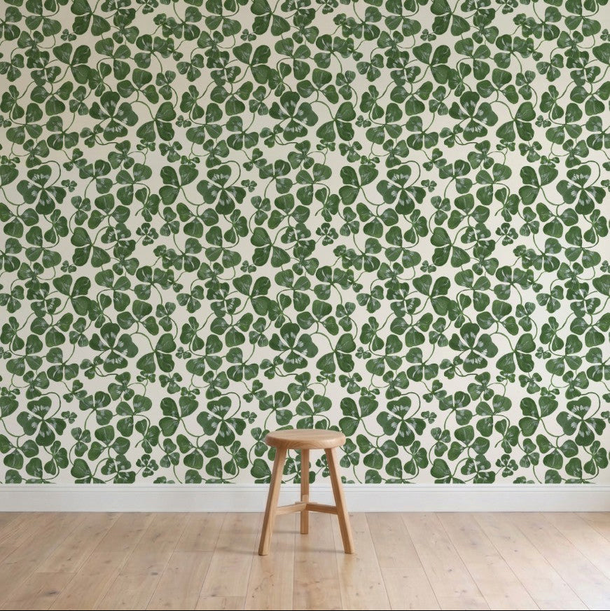 A wooden stool stands on a light wood floor in front of a wall covered with How Lucky We Are Clovers Wallpaper, featuring green leafy hand-painted designs.