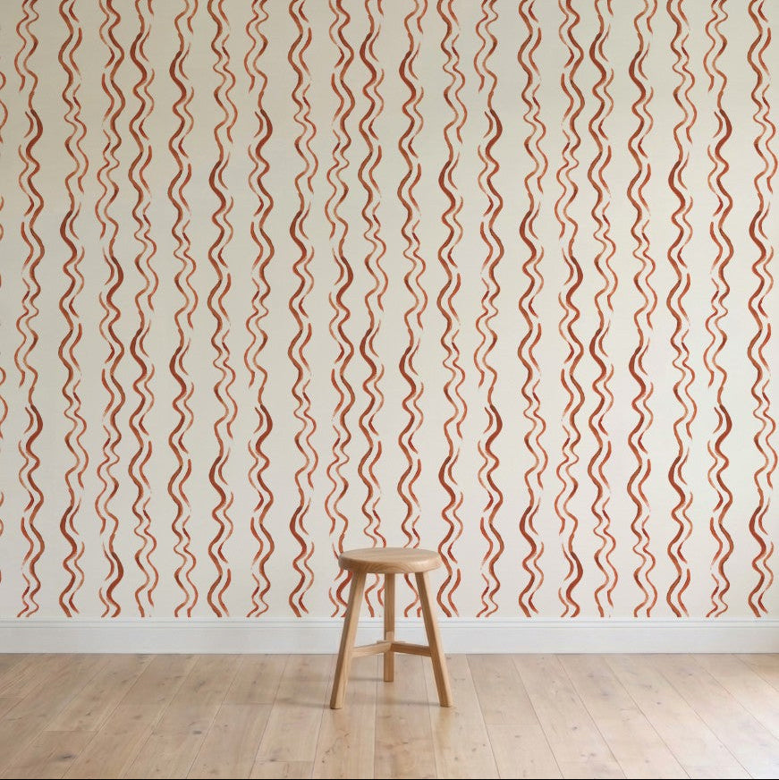A wooden stool sits on a light wood floor in front of a cream wall featuring Wiggle Stripe Wallpaper, Red—vertical wavy red and beige stripes. The minimal, well-lit setting highlights this stylish children’s wallpaper.