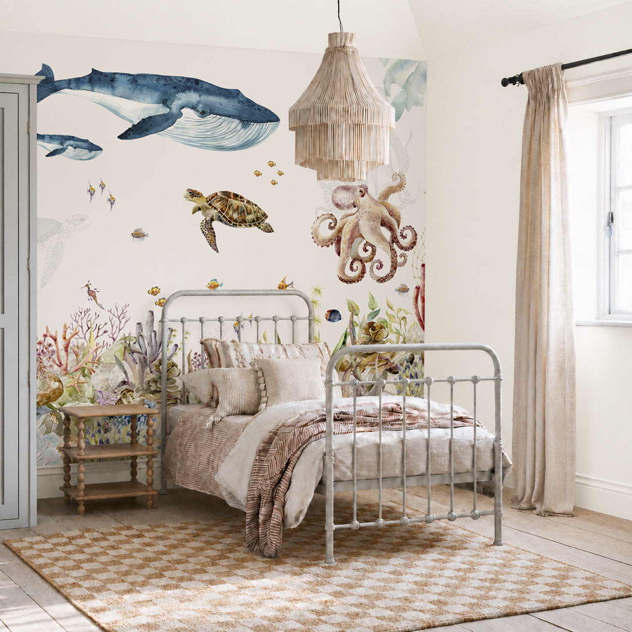 A cozy bedroom features a metal bed and woven rug, with light streaming through beige curtains to illuminate the Under The Sea Mural, Watercolour. This stunning wallpaper displays a captivating ocean scene complete with a whale, turtle, octopus, fish, and coral, bringing an enchanting under-the-sea feel to the room.