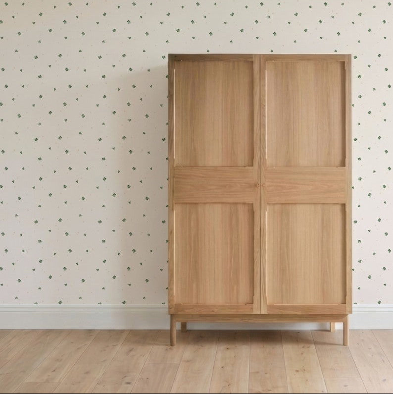 The Thank My Lucky Stars Wallpaper Sample adds a delicate touch, with its cream color and subtle green floral pattern, enhancing the quality and charm of the space behind a simple wooden wardrobe on a light wood floor.