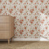 A light wooden three-drawer dresser stands on a woven rug by a wall adorned with Wild Rose Dreams Wallpaper Repeat Pattern, featuring vivid red and orange flowers with green leaves. The scene feels simple, cozy, and vintage.