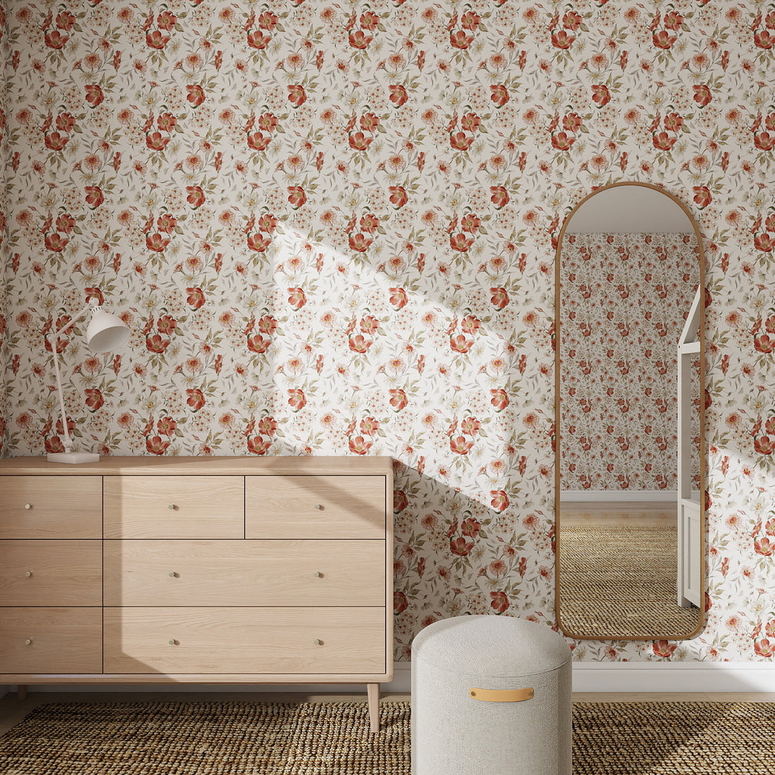 A room features Wild Rose Dreams Wallpaper Repeat Pattern, a light wood dresser with a white lamp, a round ottoman, and a tall arched mirror reflecting the wallpaper and dresser. Sunlight casts gentle shadows across the wall.