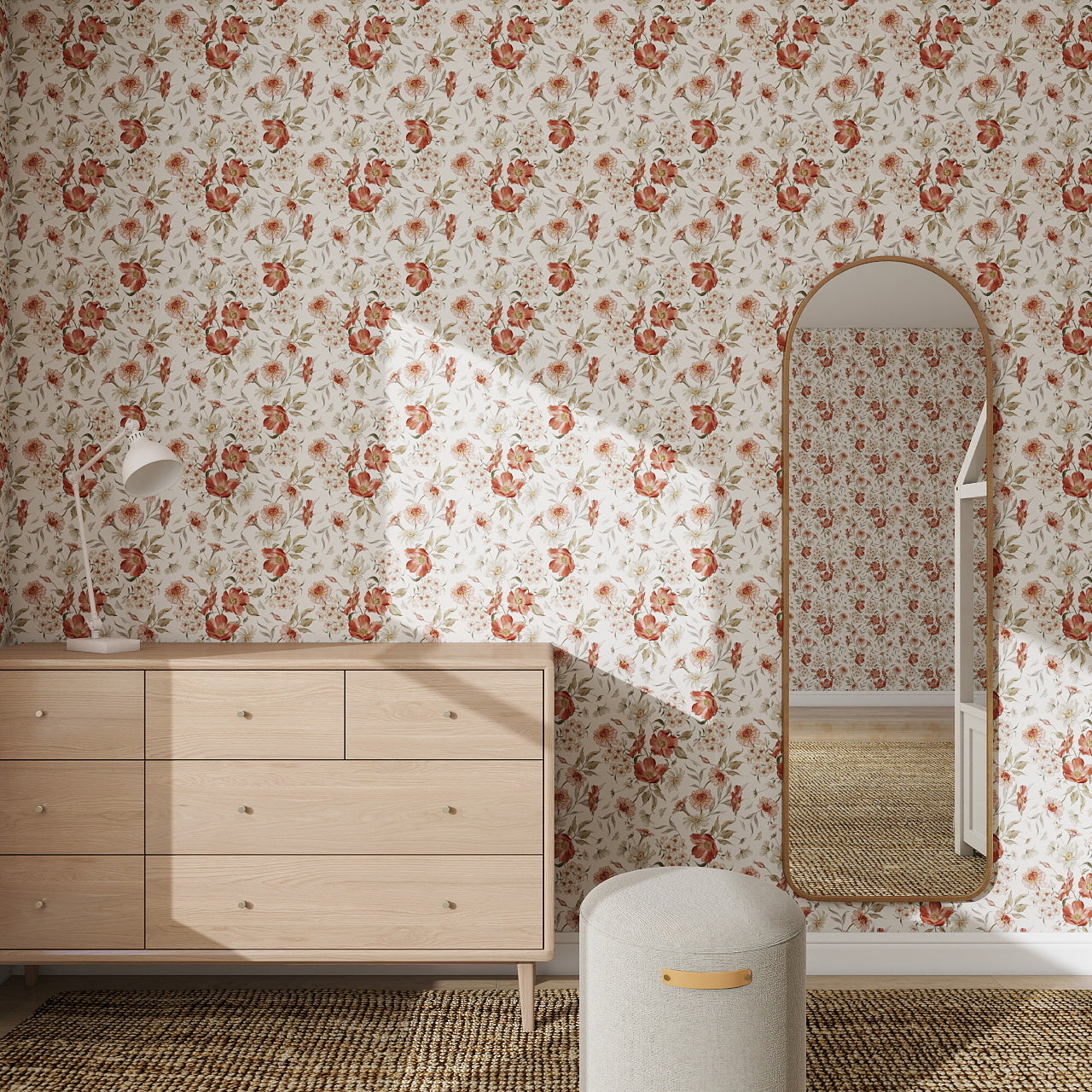 A room features Wild Rose Dreams Wallpaper Repeat Pattern, a light wood dresser with a white lamp, a round ottoman, and a tall arched mirror reflecting the wallpaper and dresser. Sunlight casts gentle shadows across the wall.