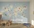 A childrens playroom for a young explorer, with the Character World Map Wallpaper Mural | Blue, illustrated animals and landmarks. A wooden table, chairs, lamp, and blue floor cushion sit on a woven rug.