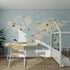 A children’s bedroom with light blue walls, a Character World Map Wallpaper Mural | Blue featuring animal illustrations, a wooden desk and chair, potted plant, white bed with brown bedding—sunlight streams in for your little adventurer.