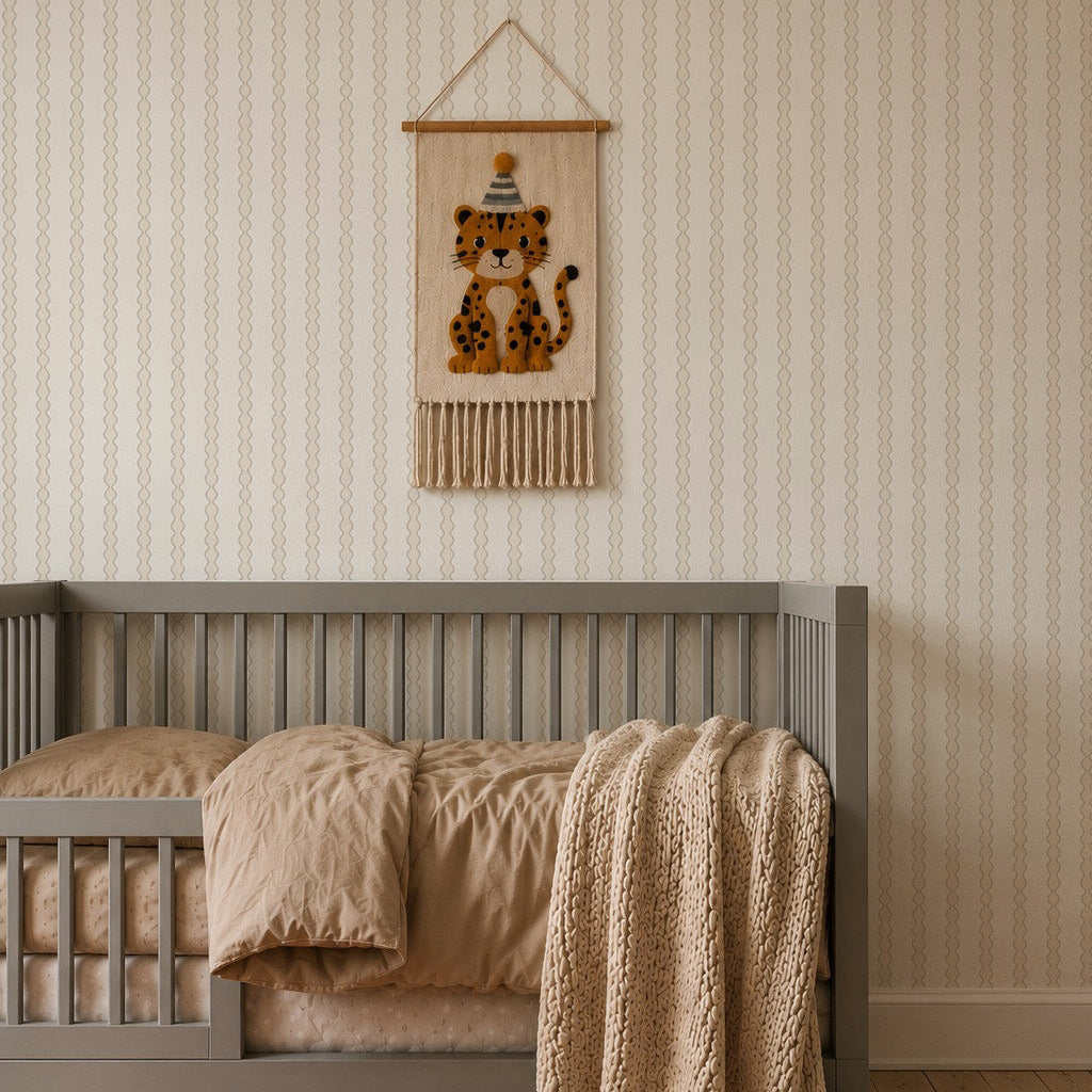 A cozy kids room features a gray crib, tan bedding, a knit blanket, a burnt orange rug, stacking toys, and charming Little Abacus Stripe Wallpaper in Off White and Taupe above the crib for a sweet finishing touch.