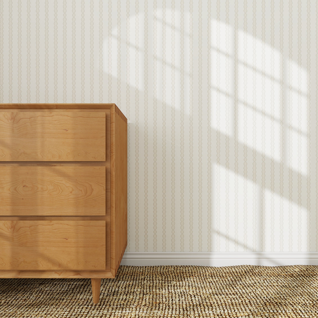 A wooden dresser stands on a woven rug beside a wall adorned with Little Abacus Stripe Wallpaper Sample in Off White and Taupe, as sunlight streams through the window, casting grid-like shadows on the elegant wallpaper.