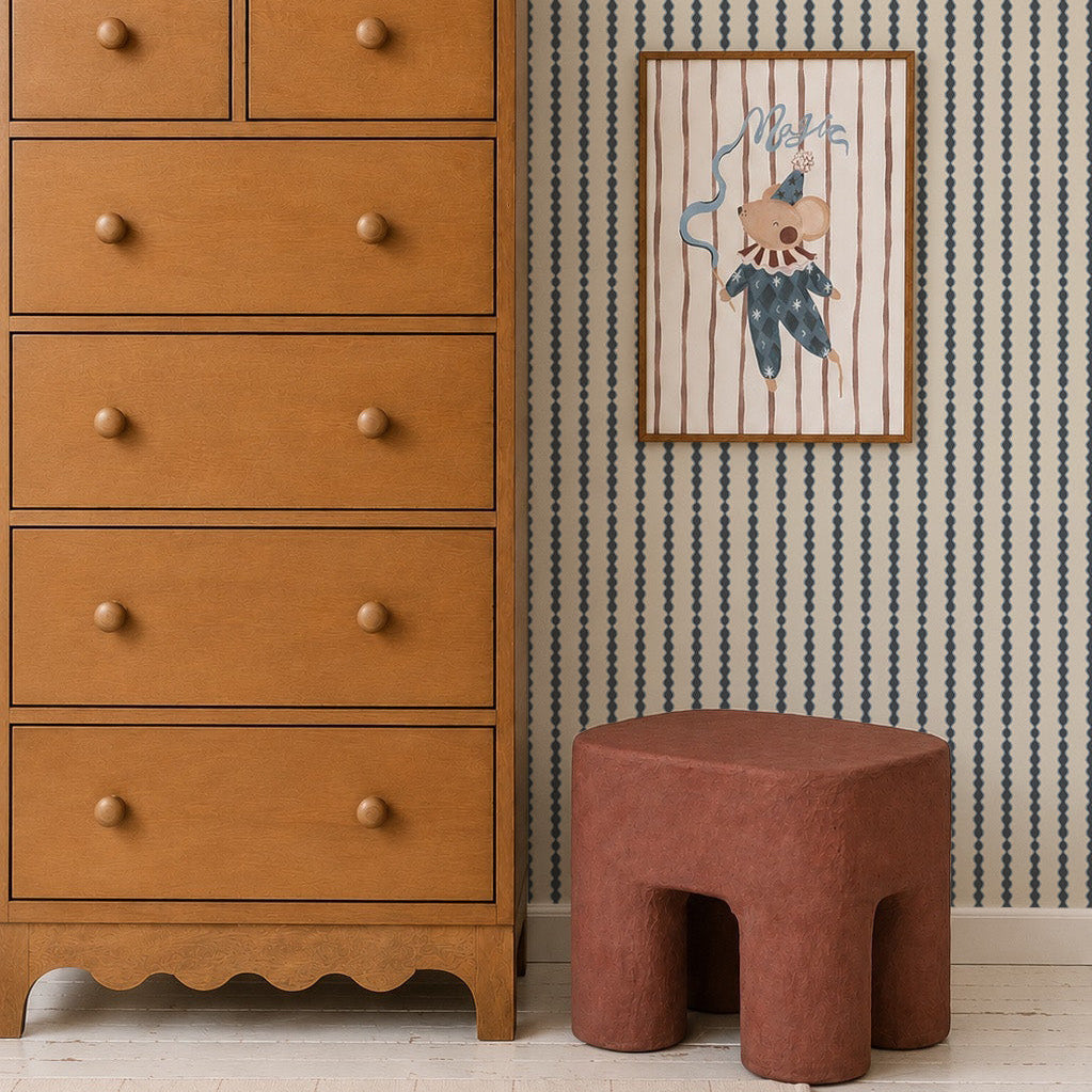 A wooden dresser stands next to a red-brown stool, with a framed animal illustration on the wall above featuring Little Abacus Stripe Wallpaper in Denim. Timeless nursery decor completes the scene, including a white grid-pattern rug and children&