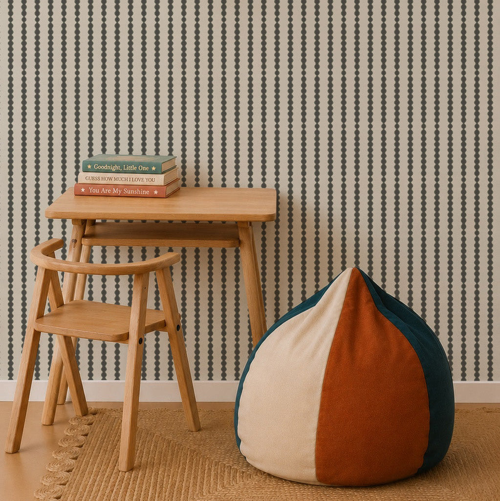 A minimalist room features a small wooden desk and chair, a stack of books, a round color-blocked bean bag, a woven rug, a pendant lamp, nursery decor for a calming vibe, and Little Abacus Stripe Wallpaper in Forest with vertical stripes on the wall.