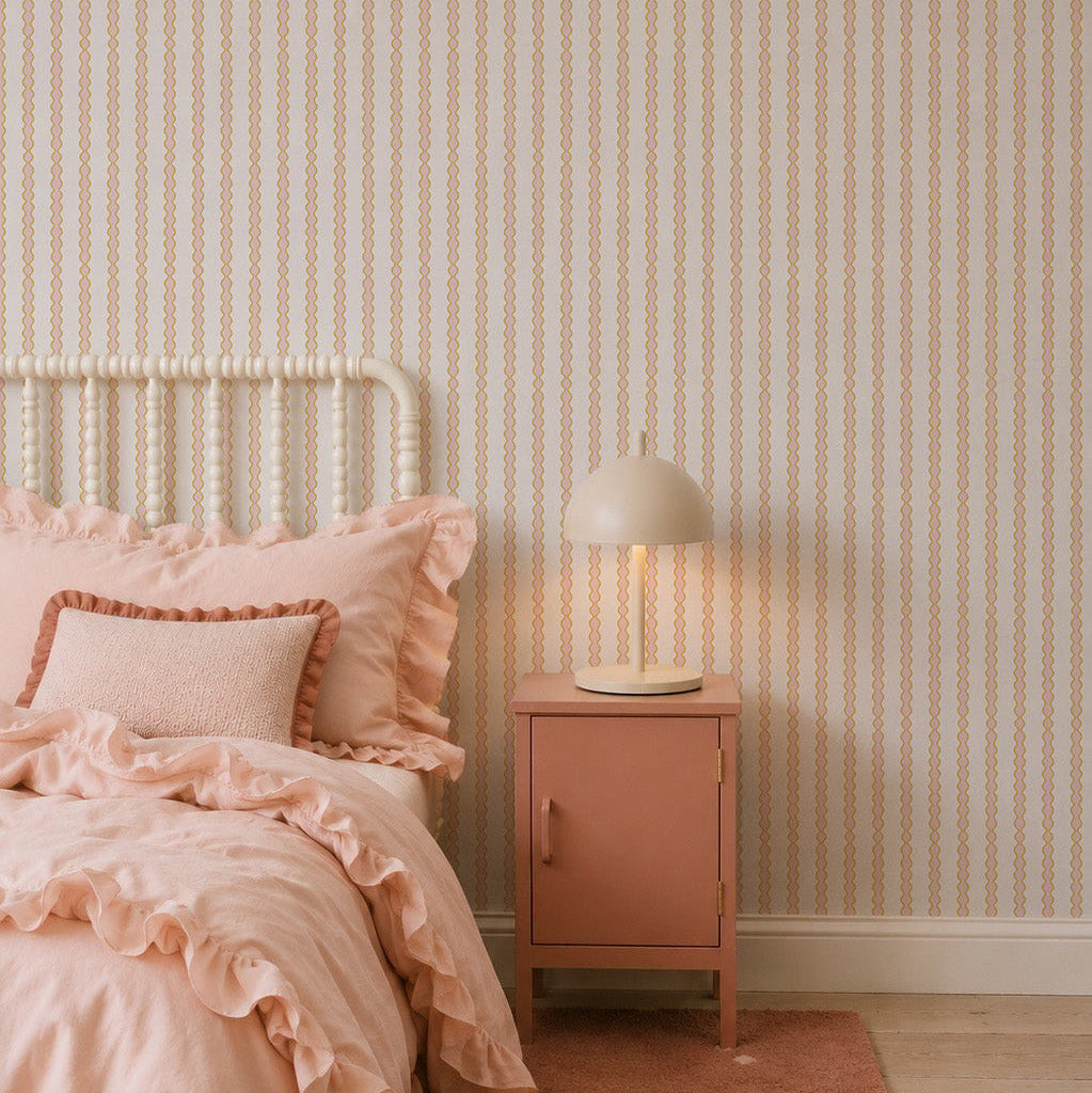 A cozy bedroom with a white spindle bed, pink ruffled bedding, pink nightstand, and beige lamp features the Little Abacus Stripe Wallpaper, Pink and Yellow—perfect nursery wallpaper for fans of soft pastel colors.