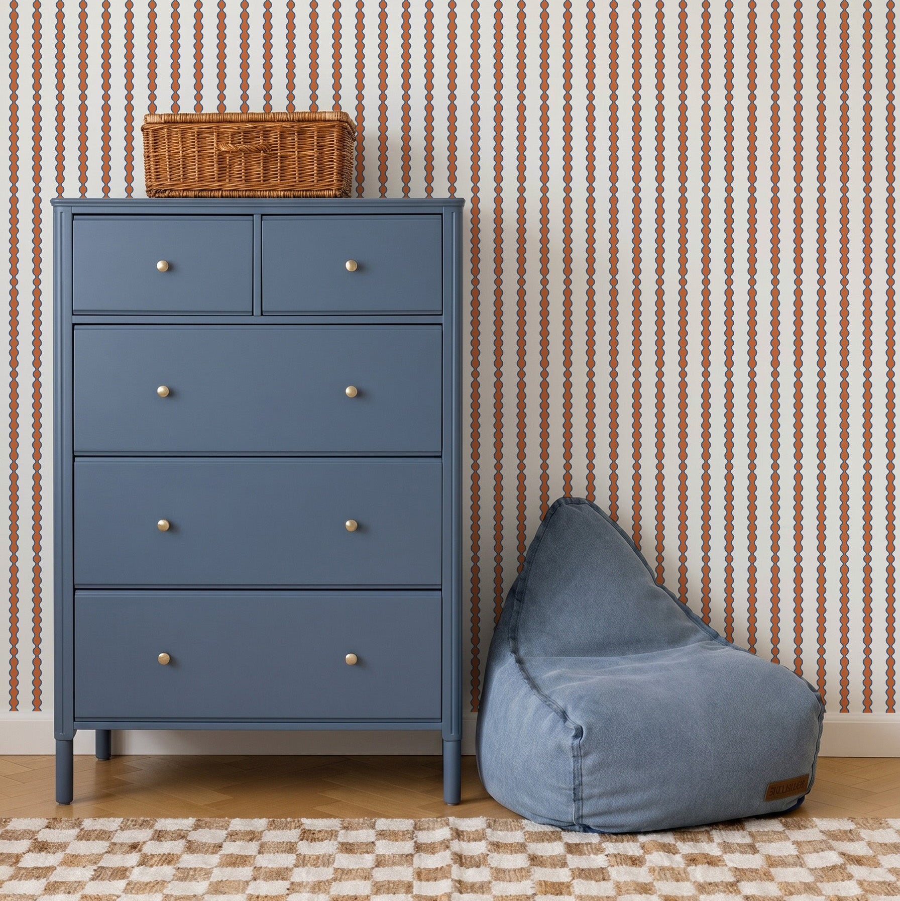 A blue dresser with silver knobs stands beside a blue bean bag chair, topped with a small wicker basket. The room features Little Abacus Stripe Wallpaper in red and blue and a checkered rug on wooden floors.