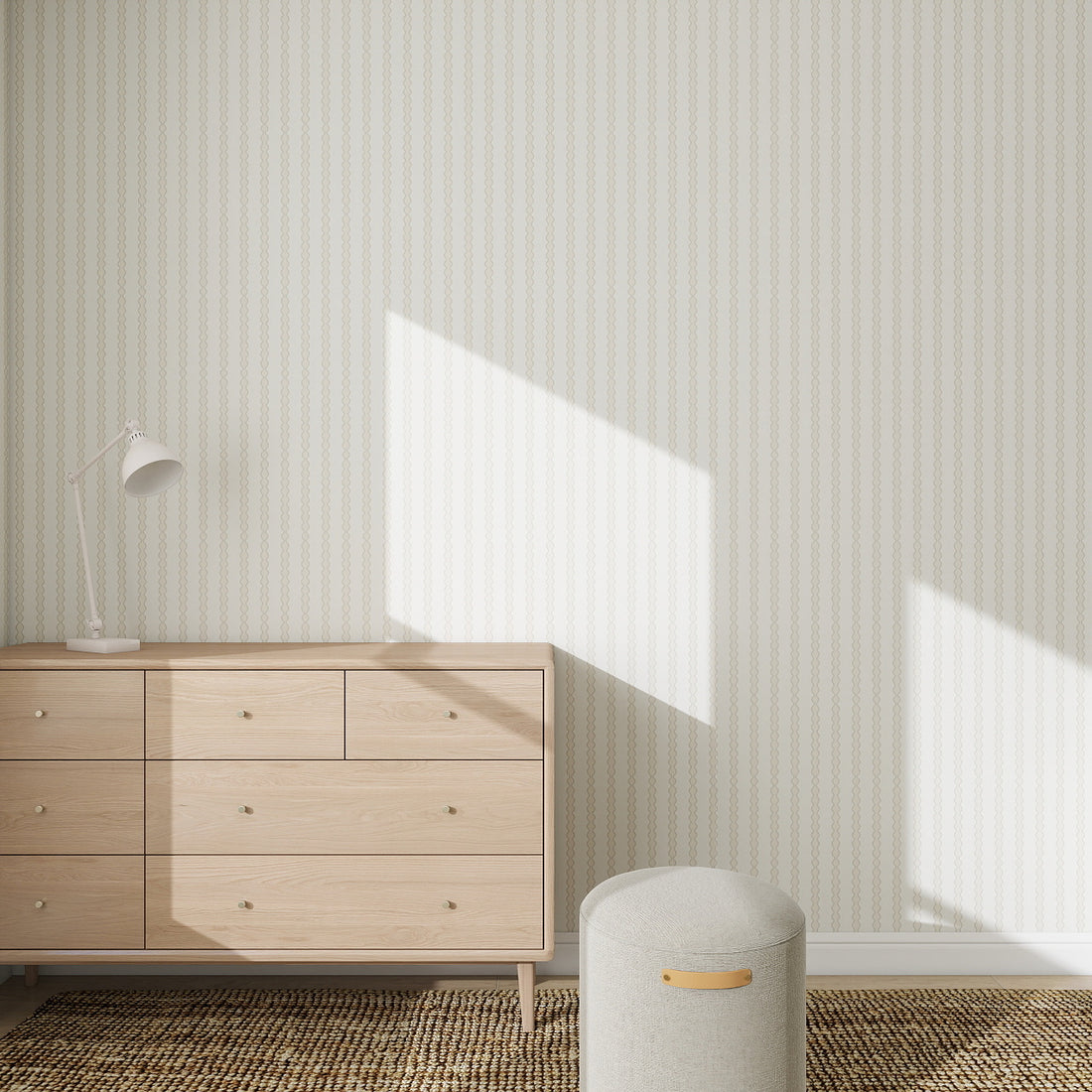 Minimalist room with a light wooden dresser, white desk lamp, and gray ottoman. Sunlight highlights the Little Abacus Stripe Wallpaper Sample in Off White and Taupe with its subtle vertical pattern. The floor features a textured carpet.