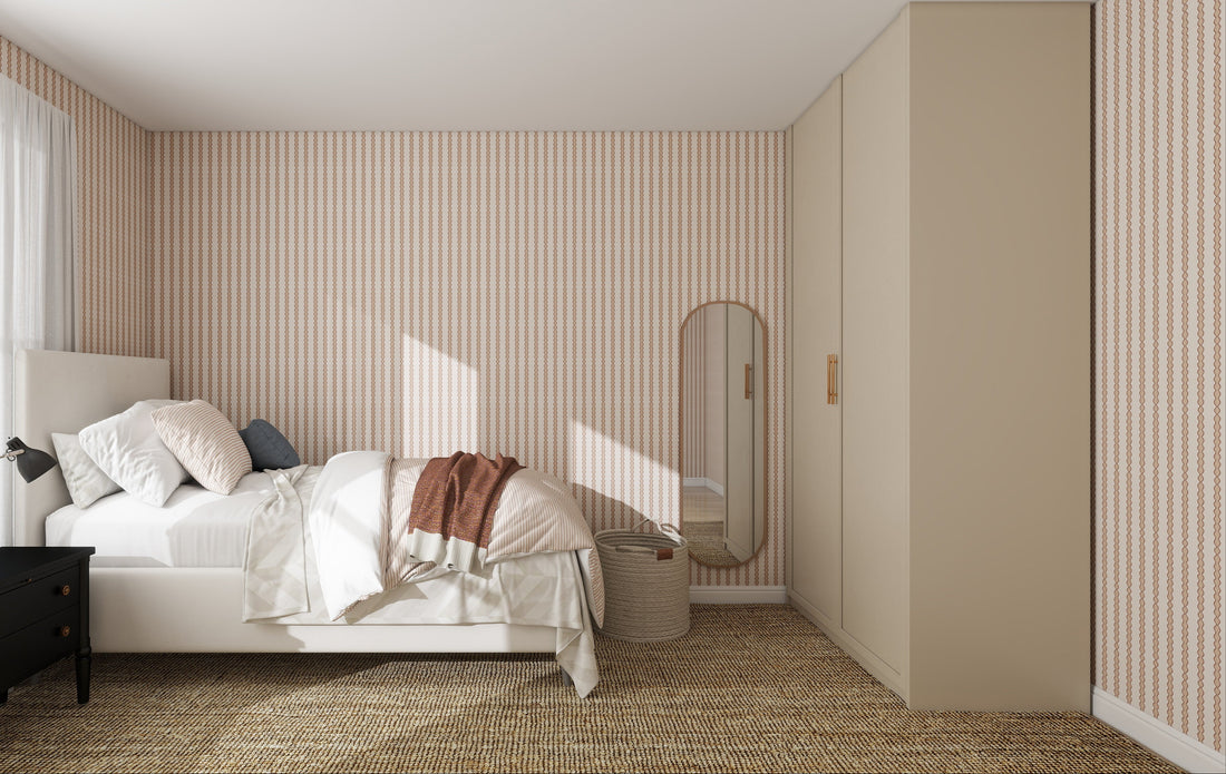 A cozy bedroom features Little Abacus Stripe Wallpaper in Rose, an unmade bed with white and beige bedding, a woven rug, black nightstand, standing mirror, laundry basket, beige wardrobe, and sunlight streaming through the window.