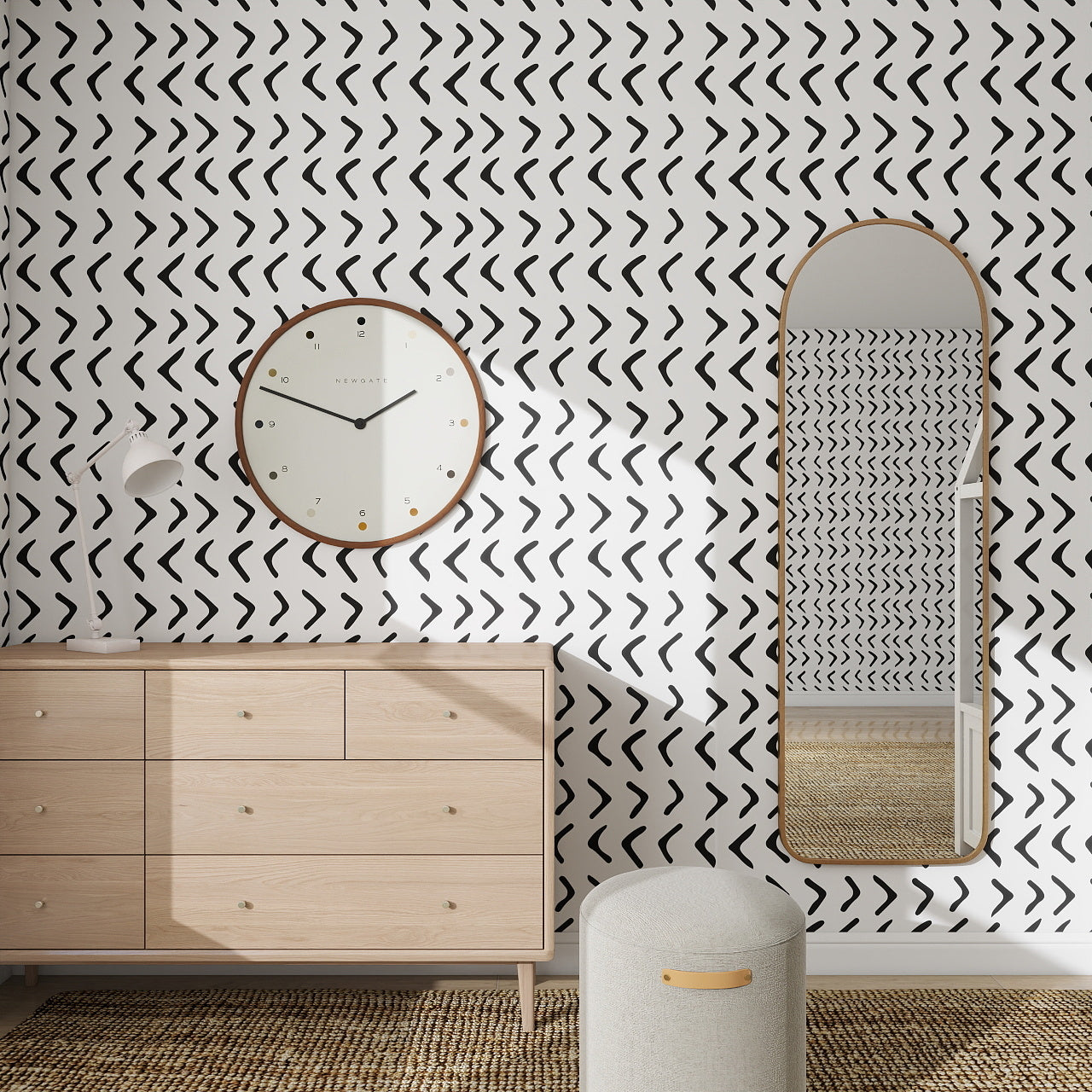 A modern room with a light wood dresser, arched mirror, round wall clock, and an accent wall featuring Andy Arrow Wallpaper Repeat Pattern in Charcoal. A textured ottoman sits atop a woven rug for added texture and style.
