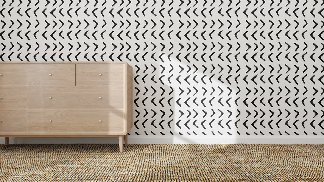A light wooden dresser stands against a white wall decorated with Andy Arrow Wallpaper Repeat Pattern in Charcoal. Sunlight casts shadows while a textured beige carpet covers the floor, creating a distinctive atmosphere.
