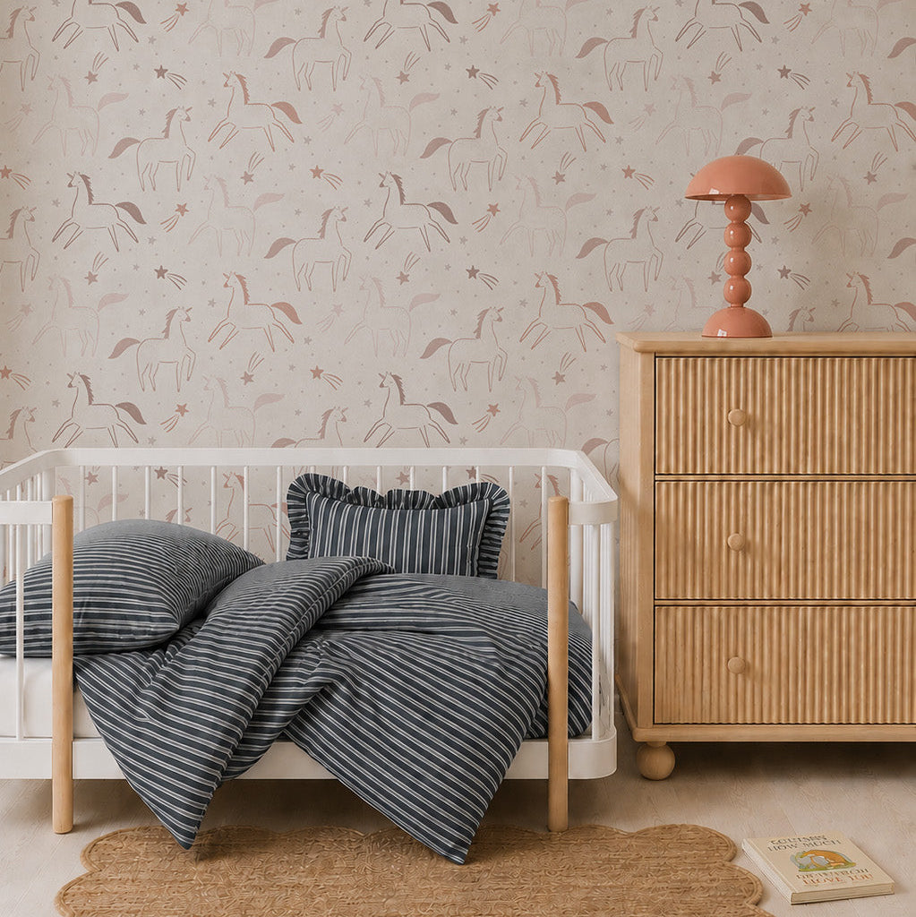 A cozy nursery features a white crib with navy striped bedding, a scalloped jute rug, a rattan dresser with a coral lamp, and whimsical Astrid Unicorn Wallpaper adorned with abstract animal and nature motifs. A book lies on the floor nearby.