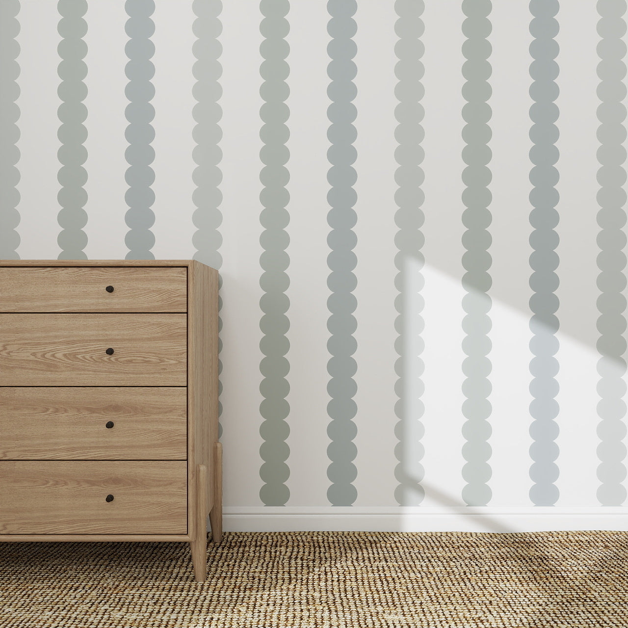 A wooden dresser stands on a beige rug next to a white wall decorated with the Scallop Stripe Wallpaper Ombre Sage and Blue Large sample. Overlapping gray circles create subtle stripes as sunlight brightens part of the wallpaper and floor.